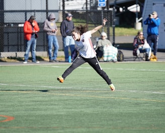 A person is mid-air on a sports field, appearing to be involved in an athletic activity. Several spectators are on the sidelines, some seated and others standing, with one individual capturing the event with a camera. The background includes a fence and various casual observers, some of whom are wearing winter clothing.