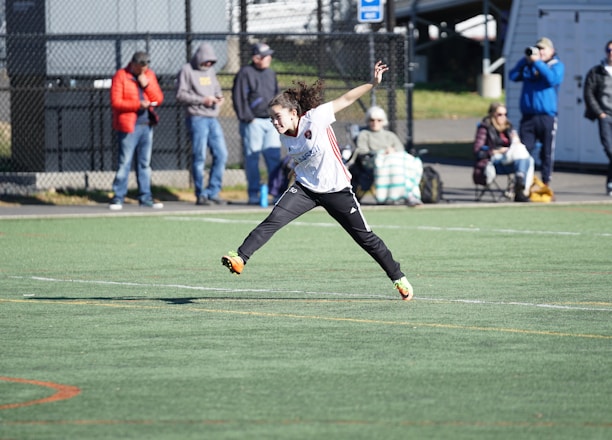 A person is mid-air on a sports field, appearing to be involved in an athletic activity. Several spectators are on the sidelines, some seated and others standing, with one individual capturing the event with a camera. The background includes a fence and various casual observers, some of whom are wearing winter clothing.