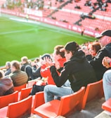 A group of people is sitting in bright red stadium seats, watching a live sports event. The stadium is well-lit with natural sunlight, and the grass on the field is vivid green. Most spectators are dressed in casual, warm clothing, reflecting cooler weather. The crowd is engaged, with many spectators clapping and attentive to the event.