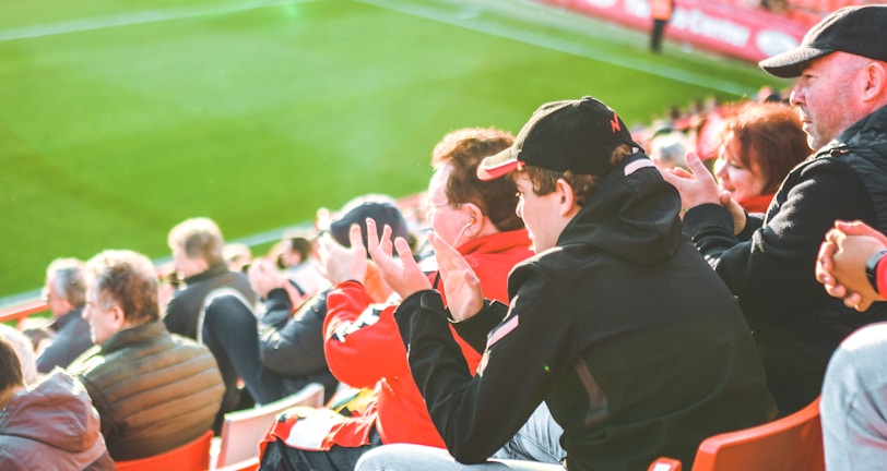 A group of people is sitting in bright red stadium seats, watching a live sports event. The stadium is well-lit with natural sunlight, and the grass on the field is vivid green. Most spectators are dressed in casual, warm clothing, reflecting cooler weather. The crowd is engaged, with many spectators clapping and attentive to the event.