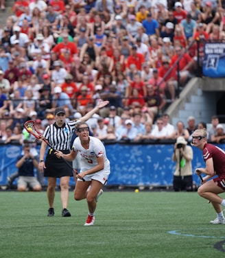 A sports event with two athletes engaged in a lacrosse game. One athlete in a white jersey is holding a lacrosse stick, appearing to prepare for a play. A referee in a striped uniform is nearby. The background features a large audience seated in a stadium, with many spectators wearing bright and casual clothing. Some photographers are also visible near the field.