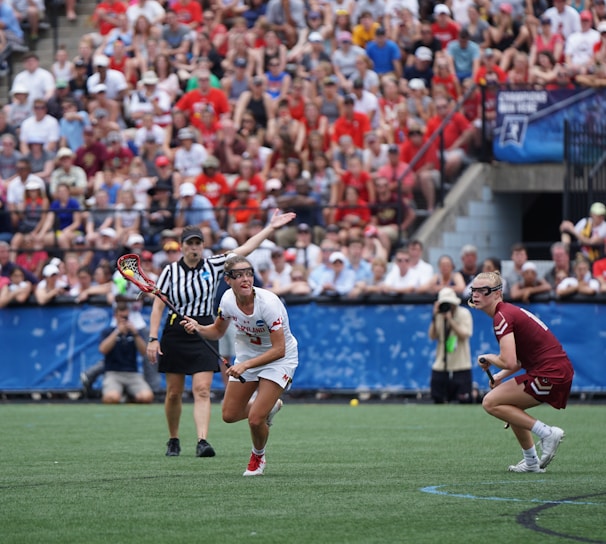 A sports event with two athletes engaged in a lacrosse game. One athlete in a white jersey is holding a lacrosse stick, appearing to prepare for a play. A referee in a striped uniform is nearby. The background features a large audience seated in a stadium, with many spectators wearing bright and casual clothing. Some photographers are also visible near the field.