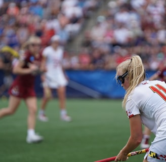 A sporting event with athletes on a grass field, one in a white jersey with the number 11 in the foreground, holding a lacrosse stick. Players in the background wear maroon uniforms. Spectators fill the stands creating a lively atmosphere.
