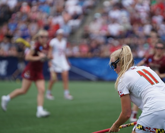 A sporting event with athletes on a grass field, one in a white jersey with the number 11 in the foreground, holding a lacrosse stick. Players in the background wear maroon uniforms. Spectators fill the stands creating a lively atmosphere.