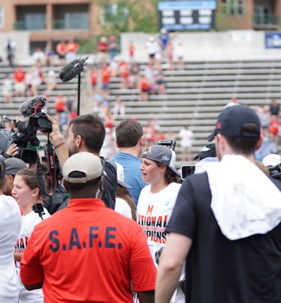A lively gathering in an outdoor sports venue with a group of people, some wearing matching shirts labeled 'National Champions.' Several individuals are holding cameras and microphones, indicating a media presence. The background shows blurred spectators sitting on bleachers.