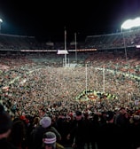 A massive crowd fills a sports stadium after an event, with people covering the field and the stands partially occupied. Bright stadium lights illuminate the scene, creating a vibrant atmosphere as individuals celebrate together.