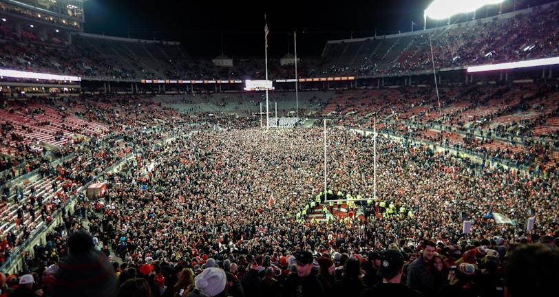 A massive crowd fills a sports stadium after an event, with people covering the field and the stands partially occupied. Bright stadium lights illuminate the scene, creating a vibrant atmosphere as individuals celebrate together.