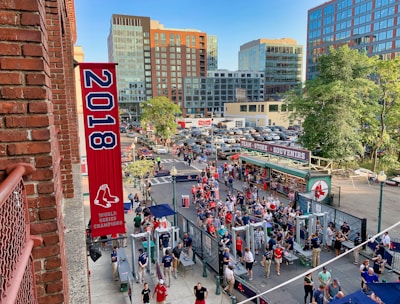 A vibrant outdoor scene featuring a large crowd of people gathered near a sports venue. A prominent red banner with '2018' and a sports logo hangs from a brick building. Nearby, there is a sign for a team store and souvenir shop. Surrounding the event are several modern high-rise buildings and a parking lot full of cars. Tall trees are visible in the background.
