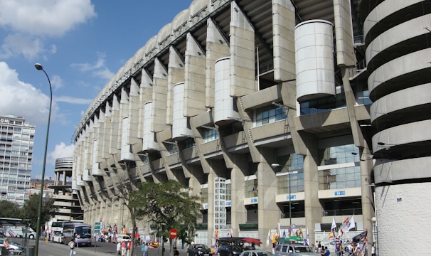 A large, modern sports stadium with a curved roof and prominent architectural features dominates the scene. Surrounding the structure, people and several vehicles are visible, indicating a busy urban environment. Flags and banners suggest an event or gathering. Nearby, urban buildings and trees add to the bustling atmosphere.
