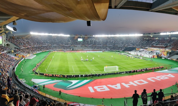 A large, brightly lit stadium filled with spectators. The field is green and well-maintained, with players actively engaged in a soccer match. The stands are crowded, and banners with writing are visible along the perimeter of the field. The atmosphere suggests excitement and energy.