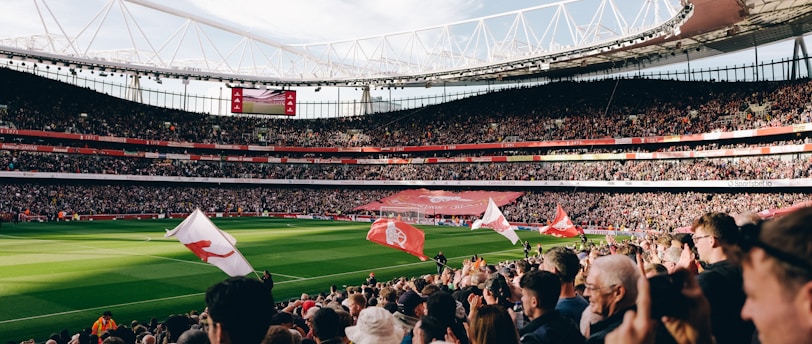 A large football stadium filled with spectators cheering and waving flags. The field is green and well-maintained, and the stands are packed with people. The atmosphere appears vibrant and energetic as people clap and celebrate.