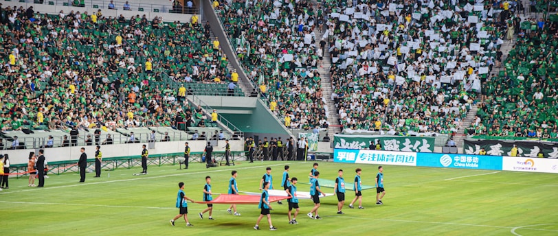 A large sports stadium is filled with spectators, the majority wearing green shirts, creating a vibrant and energetic atmosphere. In the foreground, a group of individuals dressed in blue shirts are carrying a large flag across the field. Stadium staff and officials are positioned near the field and among the crowd.