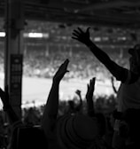 A black and white image of an enthusiastic crowd at a sports event, with individuals raising their arms in celebration or anticipation. The silhouetted figures are in the foreground, while a bright, blurred field with players can be seen in the background.
