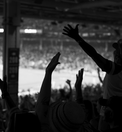 A black and white image of an enthusiastic crowd at a sports event, with individuals raising their arms in celebration or anticipation. The silhouetted figures are in the foreground, while a bright, blurred field with players can be seen in the background.
