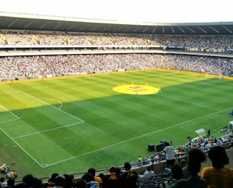 A large soccer stadium filled with a cheering crowd. The field is well-maintained with vibrant green grass, and players can be seen scattered on the pitch. The atmosphere is lively and bustling, typical of a sports event.