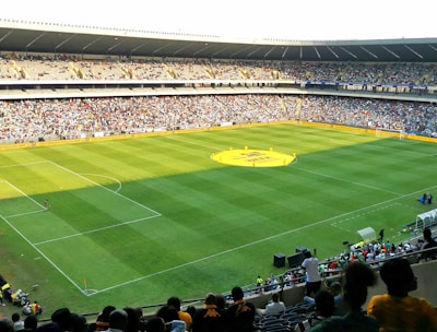 A large soccer stadium filled with a cheering crowd. The field is well-maintained with vibrant green grass, and players can be seen scattered on the pitch. The atmosphere is lively and bustling, typical of a sports event.