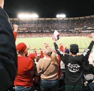 A large crowd of spectators is in a stadium during a sporting event, with many people raising their arms in excitement and wearing predominantly red clothing. The field is brightly lit and visible in the background, indicating an active scene.