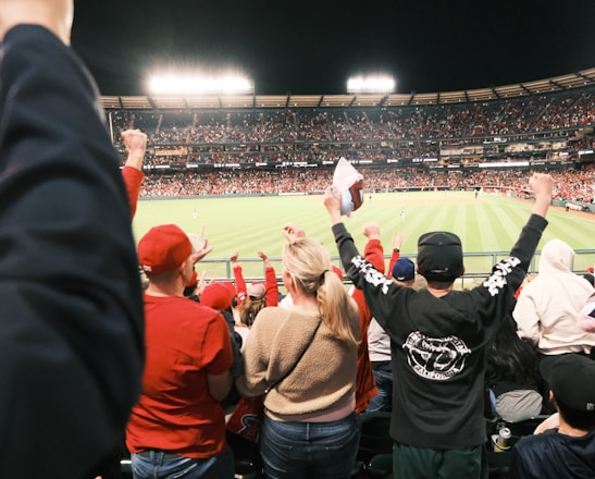 A large crowd of spectators is in a stadium during a sporting event, with many people raising their arms in excitement and wearing predominantly red clothing. The field is brightly lit and visible in the background, indicating an active scene.